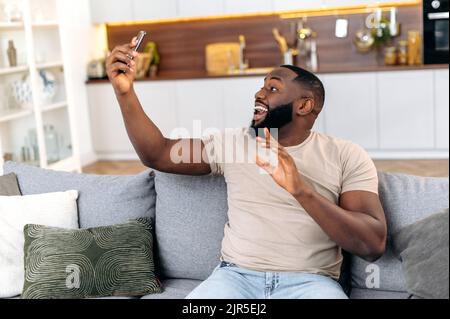 Fröhlicher afroamerikanischer junger stilvoller Mann, der im Wohnzimmer auf dem Sofa sitzt, sein Smartphone benutzt und bei einem Videoanruf spricht. Positive dunkelhäutige Videoblogger, macht ein Selfie-Handy, zeichnet einen Stream auf Stockfoto