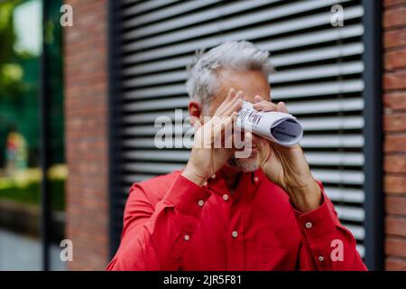 Reifer Geschäftsmann im roten Hemd, der durch die rollende Zeitung schaut und auf der Straße steht. Stockfoto