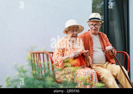 Ältere Frau, die mit ihrem Mann im Garten auf der Bank sitzt und einen roten Schal strickt. Stockfoto