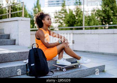 Multirassisches Teenager-Mädchen mit Rucksack und Skateboard, das während des Sommers in der Stadt sitzt. Stockfoto