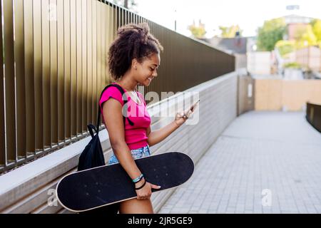 Multirassisches Teenager-Mädchen mit Rucksack und Skateboard, während des Sommers in der Stadt zu Fuß. Stockfoto