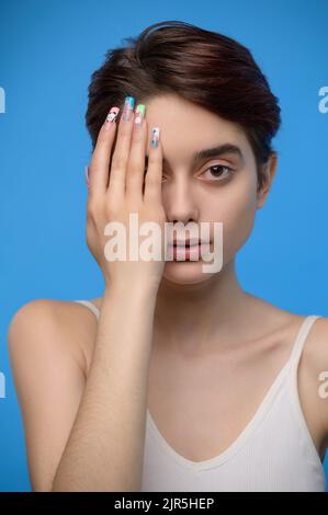 Skinny Teenager-Mädchen nettes Mädchen versteckt rechte Hälfte ihres Gesichts mit der Hand, auf blauem Hintergrund Stockfoto