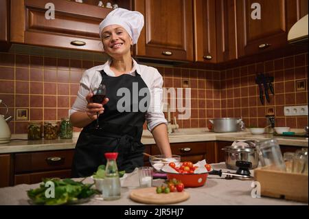 Charmante Frau, Hausfrau mit schwarzer Schürze und weißer Kochmütze, die beim Kochen in der heimischen Küche ein Glas Rotwein nippt Stockfoto