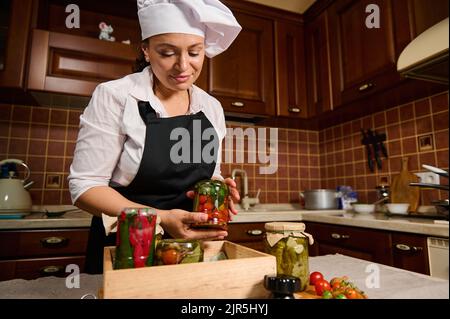 Charmante Frau mit weißer Kochmütze und schwarzer Küchenschürze hält ein Glas mit frischen Bio-Kirschtomaten aus der Dose Stockfoto