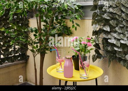 Ecke einer Terrasse mit künstlichen dekorativen Pflanzen, gelbem Metall Beistelltisch mit Gläsern und Erdbeersaft Stockfoto