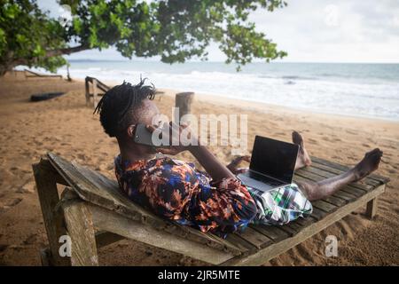 Der digitale Urlauber, der auf der Bank am Meer liegt, ruft an, während er seinen PC benutzt. Stockfoto