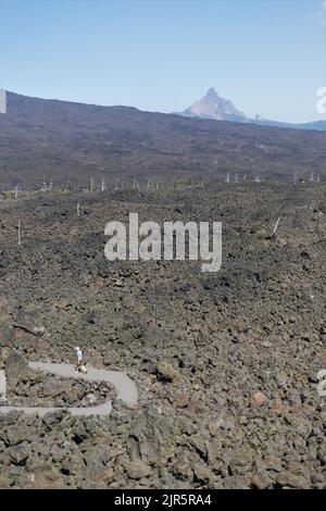 Eine Person, die auf einem Pfad durch den Lavastrom geht, mit einem Gipfel in der Ferne, wie vom Dee Wright Observatory in Oregon, USA, aus gesehen. Stockfoto