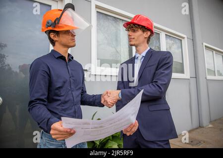 Zwei Ingenieure schütteln sich die Hände, während sie während der Bauarbeiten einen Grundriß betrachten Stockfoto