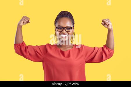 Fröhliche dunkelhäutige Frau, die Muskeln auf ihren Armen isoliert auf gelbem Hintergrund zeigt. Stockfoto