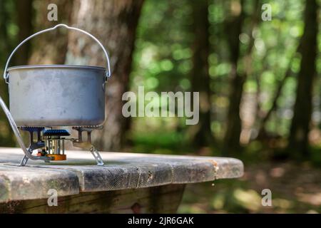 Ein Titantopf zur Herstellung von Lebensmitteln auf einem Gasherd auf einem Campingtisch aus Holz im Wald. Stockfoto