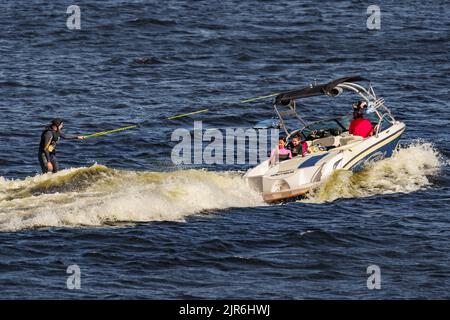 Russland, St. Petersburg, 29. Juli 2022: Mann auf Wakeboard auf Welle des Motorbootes im Sommerfluss, Wakesurfen auf Fluss Sommerferien Stockfoto