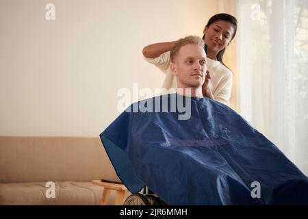 Lächelnde weibliche Betreuerin, die Haare eines jungen Mannes mit Behinderung schneidet, der im Rollstuhl sitzt Stockfoto