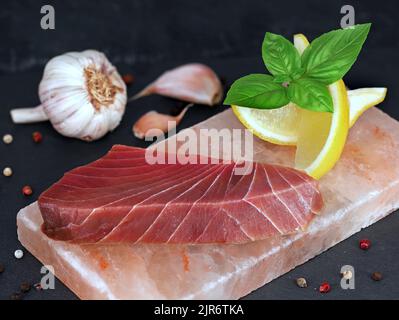 Rohes Thunfischsteak auf Salzstein mit Basilikum, Zitrone und Knoblauch auf schwarzem Schiefergrund Stockfoto