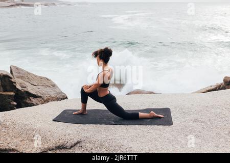 Schlanke Frau, die ihre Beine auf einer Yogamatte streckt. Frau, die Yoga am Ozean praktiziert. Stockfoto
