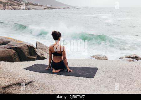 Rückansicht einer jungen Frau, die in Vajrasana sitzt, auf einer Matte im Freien. Frau meditiert, während sie auf den Ozean schaut. Stockfoto