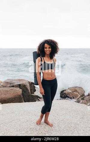 Schöne lächelnde Frau, die mit einer Yogamatte am Meer steht und die Kamera anschaut Stockfoto