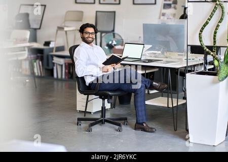 Glücklicher indischer Geschäftsmann Angestellte, der Organisator hält, der bei der Arbeit im Büro sitzt. Stockfoto
