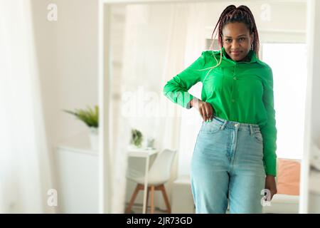 Aufgeregt fit afroamerikanische Frau nach der Gewichtsabnahme, trägt Jeans, Blick auf ihre Spiegelung im Spiegel und lächelt Stockfoto
