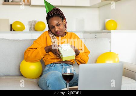 afroamerikanische Frau, die auf den Laptop-Bildschirm schaut und ein Geschenk zeigt, einen Videoanruf führt, den Geburtstag zu Hause feiert Stockfoto