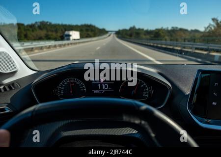 Blick des Fahrers auf den Tachometer at110 km/h oder 110 mph und die Straße verschwommen in Bewegung, Blick aus dem Inneren eines Fahrzeugs aus Sicht des Fahrers auf die Straßenlandschaft. Stockfoto