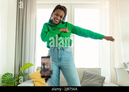 Verspielte afroamerikanische Dame, die bei der Kamera tanzt, Videos auf dem Handy zu Hause auf einem Stativ filmt und trendige Inhalte schafft Stockfoto