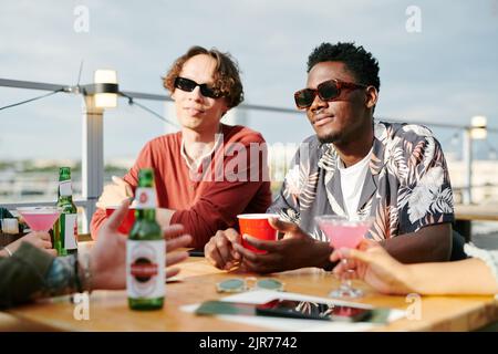 Zwei interkulturelle Männer in Sonnenbrillen und Casualwear, die im Café unter freiem Himmel Getränke trinken, während sie beim Plausch vor ihren Freunden am Tisch sitzen Stockfoto