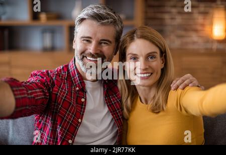 Fröhliche Männer und Frauen aus dem mittleren Alter schauen sich die Kamera an, haben Spaß und machen Fotos im Wohnzimmer Stockfoto
