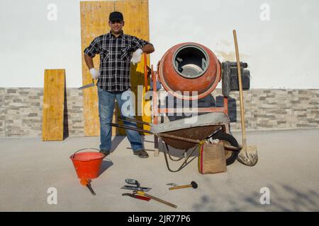 Bild eines lächelnden Bauarbeiters, der sich auf einer Baustelle gegen einen Betonmischer lehnt. Renovierung und Mauerwerk Stockfoto