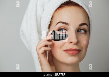 Nahaufnahme Gesicht der entzückenden jungen Frau in Handtuch nach der Dusche, mit schwarzen Kollagen Patch für die Glättung der Haut unter den Augen. Konzept-Schönheitsbehandlungen. Stockfoto