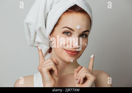 Schöne junge Frau mit Gesichtscreme und Blick auf die Kamera auf grauen Studio-Hintergrund. Angenehme Frau mit im Handtuch kümmert sich um ihre Haut. Stockfoto