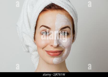 Attraktive junge Frau posiert im Studio mit weißer Maske auf der Hälfte ihres Gesichts. Kaukasische Dame im Handtuch mit Kosmetik für Anti-Aging-Verfahren zu tun. Stockfoto