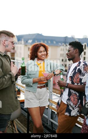 Zwei junge interkulturelle Paare in stilvoller Casualwear, die während einer Party im Freien auf der Terrasse des Dachcafés einen Drink genießen und sich unterhalten Stockfoto