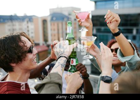 Fröhliche Männer und Mädchen, die auf der Dachterasse des Cafés im Freien auf der Dachterrasse mit Bierflaschen und Cocktails toasten Stockfoto