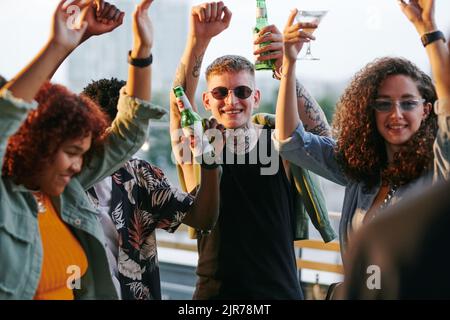 Zwei glückliche junge interkulturelle Paare, die bei einer Dachparty im Café unter freiem Himmel Alkohol tanzen und dabei die Arme hochhalten Stockfoto