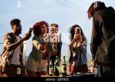 Zwei glückliche junge interkulturelle Paare mit Smartphones, die auf der Dachparty Deejay fotografieren, während sie vor ihm stehen Stockfoto