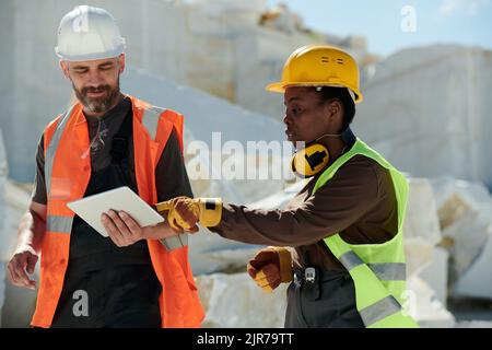 Junge, selbstbewusste Ingenieurin zeigt auf den Bildschirm der Tablette, die von einem Vorarbeiter in Schutzhelm und Arbeitskleidung während der Arbeiten im Steinbruch gehalten wird Stockfoto