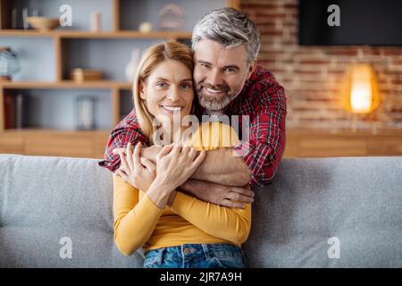 Fröhlicher, erwachsener kaukasischer Mann umarmte Frau, genoss einen zarten Moment und schaute in der Freizeit auf die Kamera Stockfoto