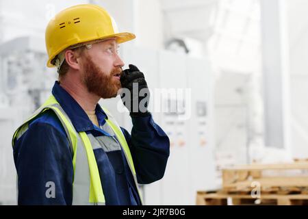 Bärtiger Arbeiter in Uniform, Hardhut und schwarzen Handschuhen mit Walkie-Talkie, während er dem Manager oder Arbeitgeber über den Produktionsprozess berichtet Stockfoto