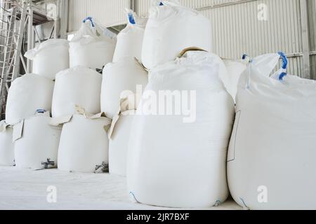 Haufen riesiger weißer Säcke mit losen Rohstoffen, die für den Produktionsprozess in einem geräumigen Lager oder Lagerraum vorbereitet werden Stockfoto