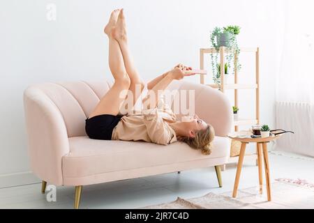 Glückliche Frau liegt auf dem Rücken auf einem beigefarbenen Sofa, die Beine nach oben, schaut auf ein digitales Tablet Stockfoto