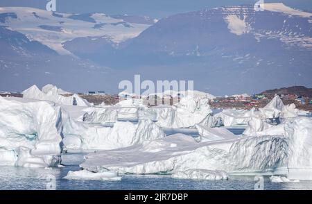 Große Eisberge vor der kleinen Gemeinde Saqqaq mit aufragenden Bergen im Hintergrund in der Disko Bay, Grönland, am 18. Juli 2022 Stockfoto