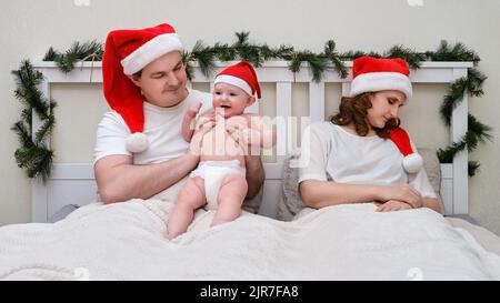 Ehemann spielt mit Kind, während Frau auf dem Bett schläft, das für weihnachten und Neujahr dekoriert ist, Familienpaar im heimischen Schlafzimmer Stockfoto