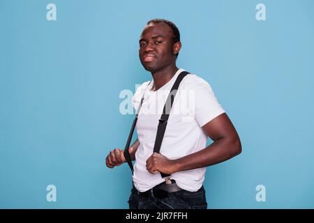 Fröhlicher junger Mann, der die Kamera anlächelt, während er an den Hosenträgern zieht. Porträt von selbstbewussten cool aussehende afrikanische Person auf blauem Hintergrund in Mode Kleidung stehen. Studioaufnahme Stockfoto