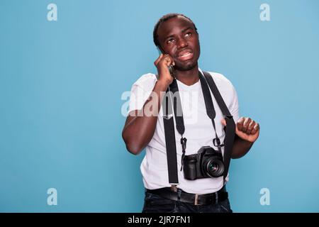 Fröhlicher Fotofan, der auf dem Mobiltelefon spricht, während er ein Fotogerät hat. Glücklicher Profi-Fotograf mit DSLR-Kamera sprechen am Telefon, während lächeln. Studioaufnahme Stockfoto
