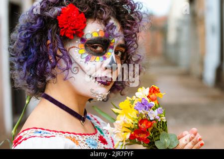 Outdoor-Porträt einer jungen lateinischen kaukasischen Frau, mit Gesicht gemalt als La Calavera catrina, stehend auf verschwommenem Friedhof im Hintergrund und Kopierraum. Stockfoto