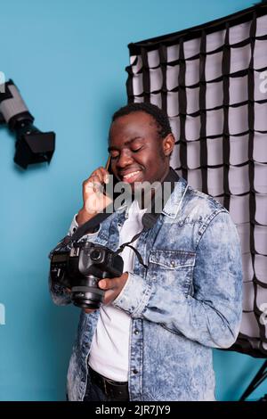 Selbstbewusster Fotofan, der in einem professionellen Produktionsstudio auf blauem Hintergrund telefoniert. Fröhlicher Fotograf, der während eines Fotoshootings auf dem Smartphone spricht. Stockfoto