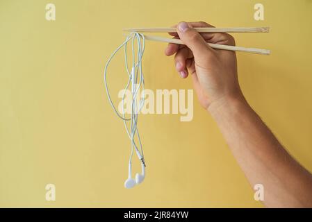 Essen und Musik. Was braucht man mehr. Beschnittene Studioaufnahme eines Mannes mit Ohrhörern, die vor einem gelben Hintergrund um Essstäbchen gewickelt sind. Stockfoto