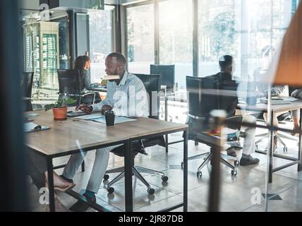 Geschäft, Büro und Arbeit mit einem Mann, der an seinem Schreibtisch in einem Glasgebäude sitzt und im Hintergrund mit Kollegen arbeitet. Ein geschäftiges Unternehmen Stockfoto