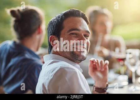 Ich bin am glücklichsten, wenn ich mit meinen Freunden bin. Porträt eines glücklichen jungen Mannes, der mit Freunden auf einer Dinnerparty im Freien eine Mahlzeit teilt. Stockfoto