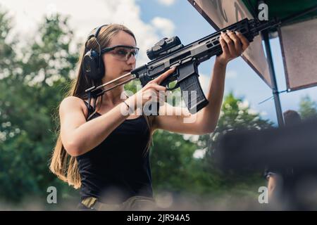 Starke, selbstbewusste langhaarige kaukasische Frau in einem schwarzen Panzer mit großem schwarzen Gewehr und Lupe, die sich auf einen Schießwettbewerb vorbereitet. Mittlere Außenaufnahme. Hochwertige Fotos Stockfoto